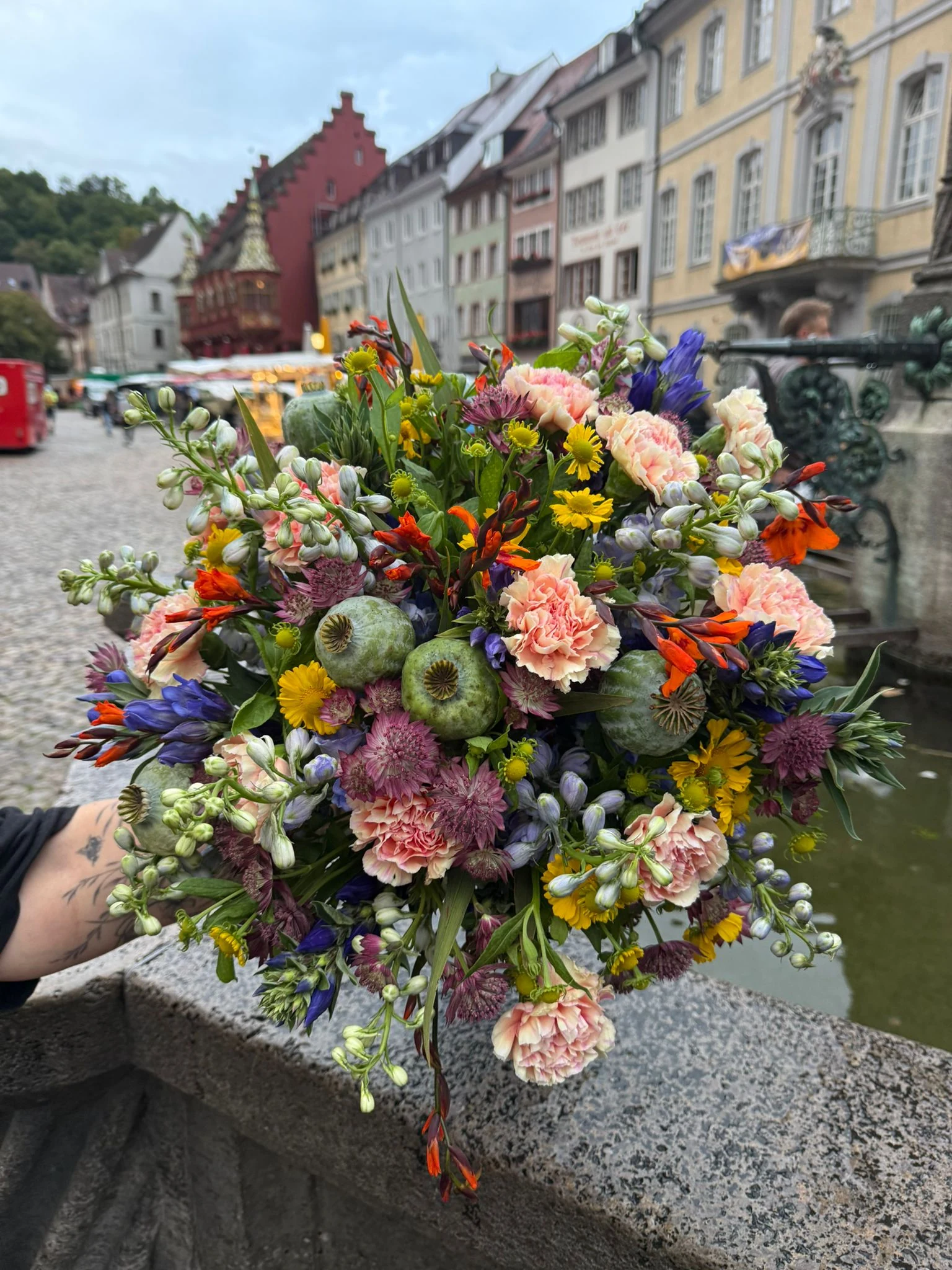 Bunter Strauß mit rosa Nelken, gelben Blüten und blauem Rittersporn vor einer Häuserzeile